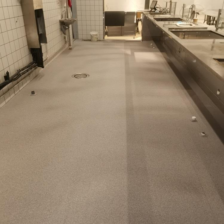 Vaaleanharmaa, täplikäs lattiapinnoitus teollisuuskeittiössä, jossa on lattiakaivo ja ruostumattomasta teräksestä valmistettuja työtasoja.