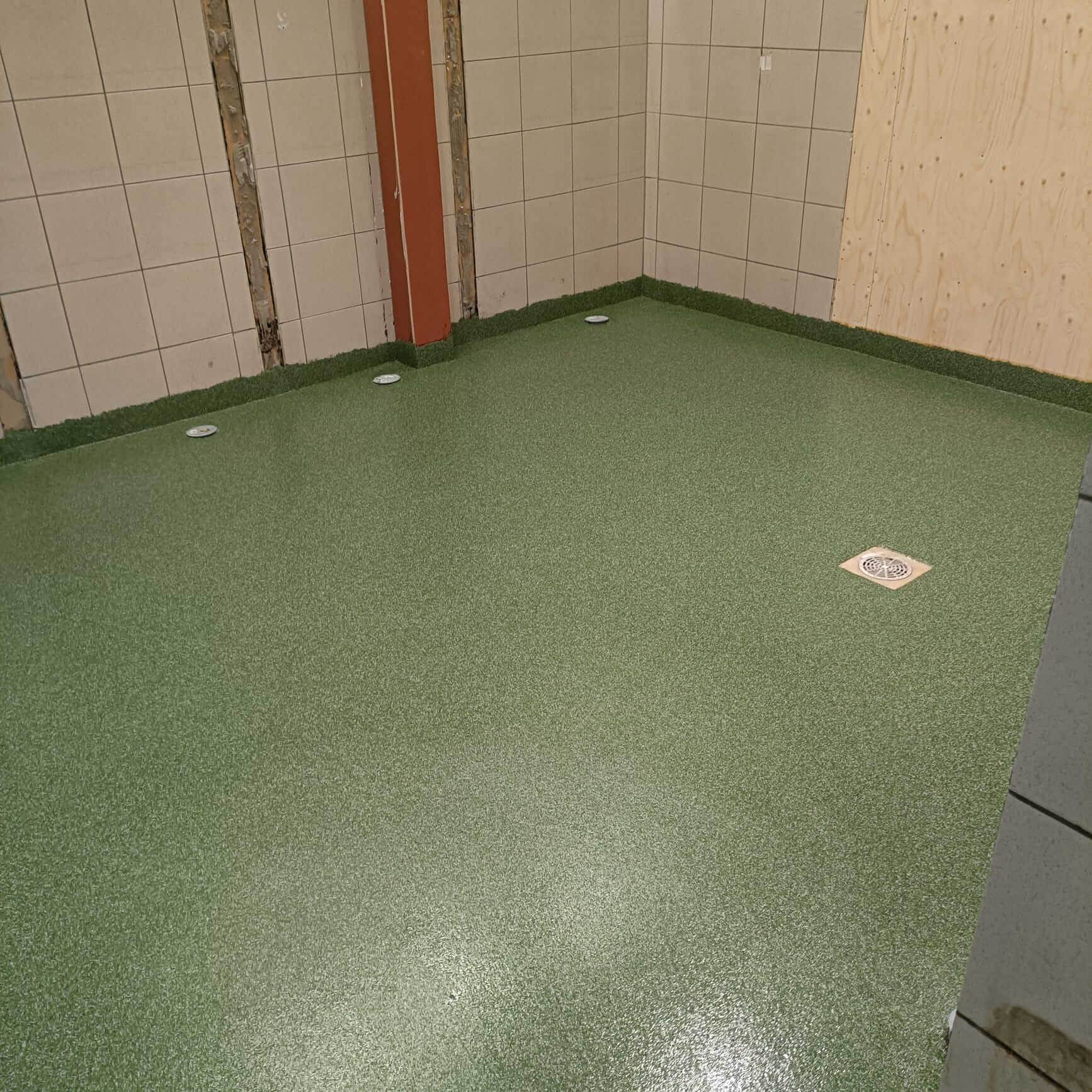 Huoneessa on vihreä, rakeinen lattiapinnoitus, jossa on lattiakaivo ja seinien alaosassa vihreä jalkalista.